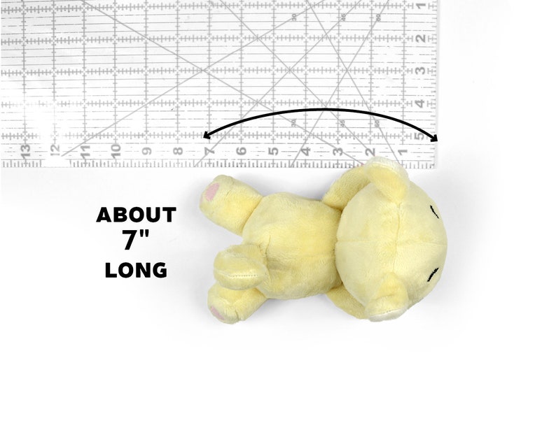 May include: A yellow plush teddy bear lying on its back, measuring approximately 7 inches long. The bear has a simple face with black eyes and a small mouth.