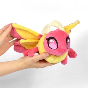 May include: A plush toy moth with a pink head and body, yellow wings with pink accents, and large, cartoonish eyes. The moth has small antennae and is held against a white background.