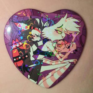 May include: Heart-shaped button featuring a cartoon illustration of three characters. The characters are in various colors, including black, white, red, and pink. The background is purple with a web-like design and holographic accents.