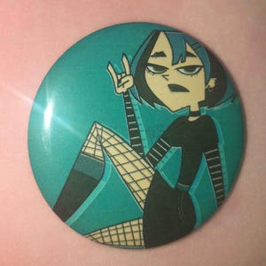Può includere: Un bottone verde acqua con un'illustrazione a cartoni animati di un personaggio con capelli neri e un abito nero. Il personaggio fa un gesto della mano rock-on. Il bottone ha una finitura lucida e uno sfondo verde acqua.