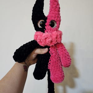 May include: A plush toy featuring a split-color design with one side in black and the other in bright pink. The creature has large, round, glittery black eyes and a textured, fuzzy yarn finish. It wears a matching pink ruffled scarf around its neck.