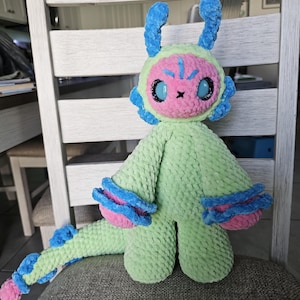 May include: A plush toy creature made of soft light green yarn with a pink face, large black eyes, and blue antennae. It features blue and pink ruffled cuffs on its arms, a long tail with blue and pink accents, and a textured, fuzzy appearance.