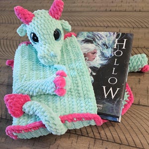 May include: A light mint green crocheted dragon plush with bright pink horns, claws, and tail tip, featuring large black eyes. The dragon is positioned next to a book titled Hollow, which displays an illustration of a person and a dragon.