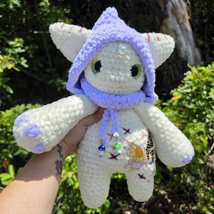 May include: A white crocheted plush creature with a lavender hood, large green eyes, and lavender paw pads. The plush features embroidered stars, a butterfly patch, and two small mushroom charms hanging from the hood strings with a silver moon charm.
