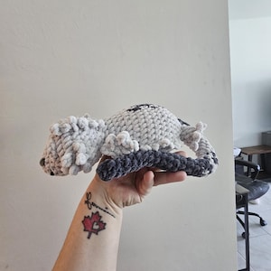 May include: A handmade crochet lizard in shades of gray and dark blue. The plush reptile has a textured body, small legs, and a curled tail. The lizard is held up against a neutral wall.