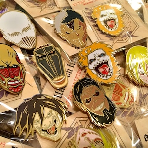 Op de afbeelding: Een verzameling emaille pins met diverse karakterontwerpen, elk met unieke gezichtsuitdrukkingen, haarkleuren en metalen omlijningen. Elke pin is afzonderlijk verpakt in een doorzichtig plastic hoesje en bevestigd op een kartonnen kaartje.