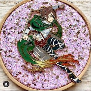 Op de afbeelding: Een metalen emaille speld met een anime-personage met bruin haar, een bril en een ooglapje, gekleed in een groene cape en uniform. Het personage staat in een dynamische houding met een zwaard en uitrusting tegen een ronde achtergrond van roze glitter.