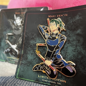 May include: A hard enamel pin featuring the character Maki Zen'in in a kneeling pose, holding a three-section staff with a gold chain. The pin is mounted on a black backing card with the text MAKI ZEN'IN and LIMITED EDITION HARD ENAMEL PIN.