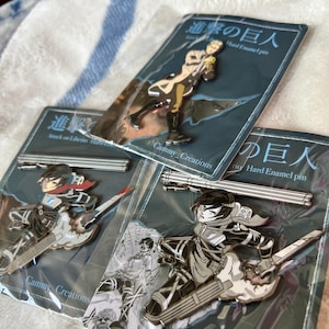 May include: Three hard enamel pins featuring anime-style characters in dynamic poses, each mounted on a branded backing card with Japanese text and the label Cammy_Creations. The pins depict figures in tactical gear with swords and equipment.