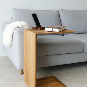 Könnte beinhalten: Ein moderner Beistelltisch aus hellem Holz mit rechteckigem Design. Die Tischplatte bietet Platz für ein Buch und ein Smartphone. Der Tisch steht neben einem grauen Sofa. Die Tischhöhe beträgt ca. 60 cm.