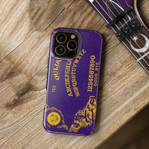 Könnte beinhalten: Lila Handyhülle mit Ouija-Brett-Design. Das Gehäuse hat goldfarbene Buchstaben und Grafiken, einschließlich des Alphabets, Zahlen und die Wörter "Ja" und "Auf Wiedersehen". Auf einer Holzoberfläche.