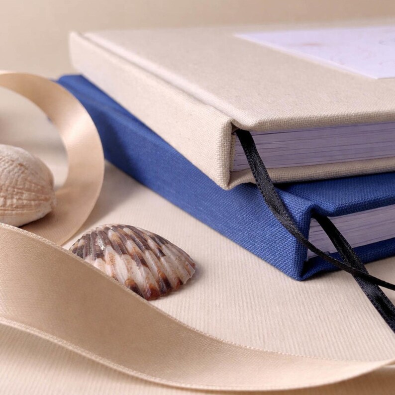May include: Two hardcover journals, one beige and one blue, are stacked. A black ribbon bookmark is visible. A beige ribbon and two seashells are in the foreground. The journals have a textured fabric cover. The image has a soft, neutral color palette.