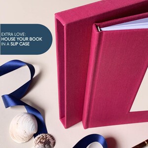 May include: A close-up of a magenta book and slipcase. The book is open, revealing white pages. A navy blue ribbon and seashells are in the foreground. Text on the left reads: "EXTRA LOVE: HOUSE YOUR BOOK IN A SLIP CASE."