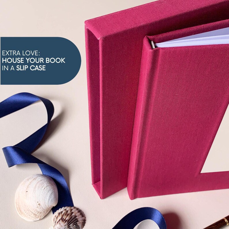 May include: A close-up of a magenta book and slipcase. The book is open, revealing white pages. A navy blue ribbon and seashells are in the foreground. Text on the left reads: "EXTRA LOVE: HOUSE YOUR BOOK IN A SLIP CASE."