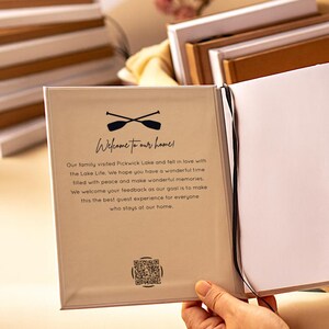 May include: A guest book with a beige cover featuring a black graphic of two crossed oars above the text Welcome to our home! followed by a printed message about Pickwick Lake and a QR code at the bottom. A thin black ribbon bookmark is attached.