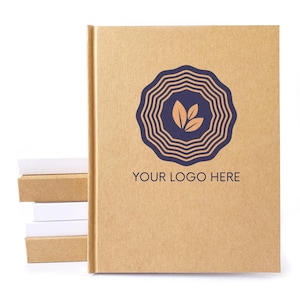 May include: A tan hardcover notebook with a dark blue circular logo featuring concentric wavy lines and three leaves in the center. The text "YOUR LOGO HERE" is printed in dark blue below the logo. A stack of small tan and white blocks sits nearby.