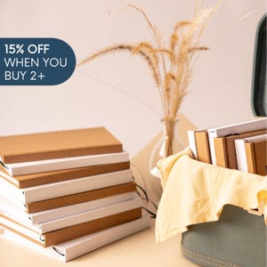 May include: A stack of rectangular notebooks with brown covers and white pages, alongside a green container holding additional notebooks and a yellow cloth. A dark blue graphic in the corner displays the text 15% OFF WHEN YOU BUY 2+.