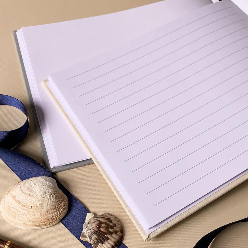 May include: Open lined journal with a light beige cover and gray spine. A dark blue ribbon and two seashells are placed near the journal. The pages are white with gray lines for writing.