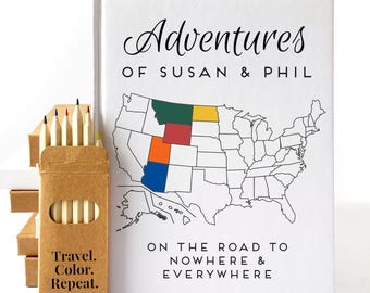 Peut inclure: Un livre blanc intitulé Adventures of Susan & Phil On the Road to Nowhere & Everywhere avec une carte en noir et blanc des États-Unis où certains États sont colorés en vert, jaune, rouge, orange et bleu, accompagné d'une boîte de crayons.