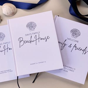 May include: Three white hardback guest books featuring dark blue text and shell illustrations. One book reads Davis Family Beach House with coordinates 39.61697 N 74.19768 W. Another reads Welcome family & friends, Sherrell's Beach Condo, Est. 2026.