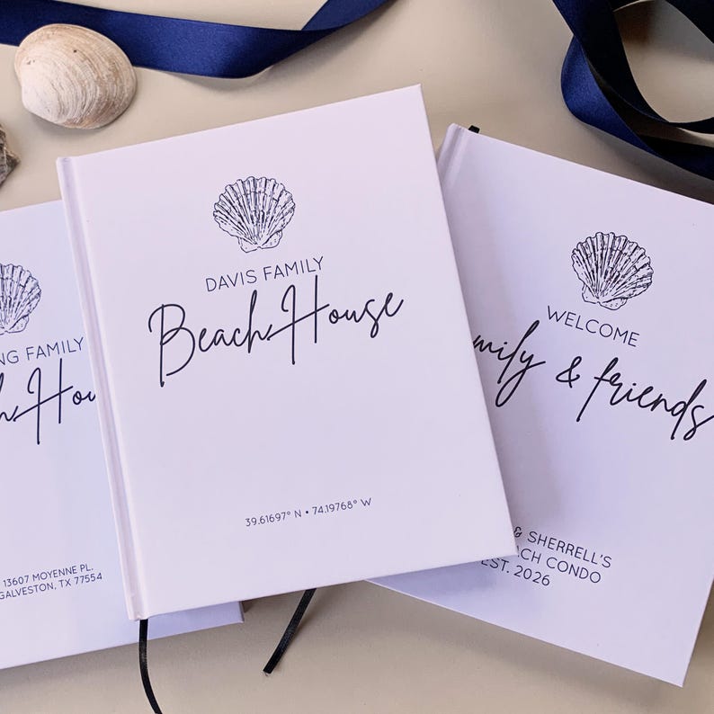May include: Three white hardback guest books featuring dark blue text and shell illustrations. One book reads Davis Family Beach House with coordinates 39.61697 N 74.19768 W. Another reads Welcome family & friends, Sherrell's Beach Condo, Est. 2026.