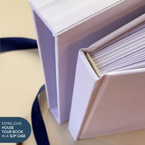 May include: A white book with a matching white slipcase, featuring a visible beige fabric spine binding and white pages. A dark blue ribbon is positioned nearby. The text on the image reads: EXTRA LOVE: HOUSE YOUR BOOK IN A SLIP CASE.