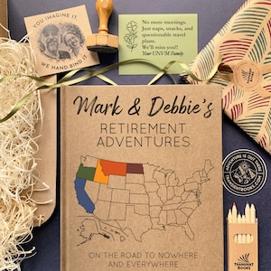 May include: A retirement adventure journal titled "Mark & Debbie's Retirement Adventures" with a map of the United States. Includes a stamp, a card with text, a wrapped gift, and a box of colored pencils. The journal's text reads "On the road to nowhere and everywhere."