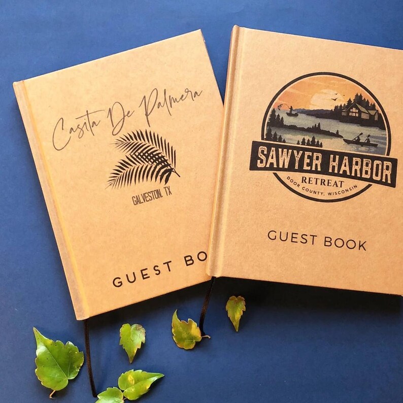 May include: Two tan hardcover guest books with black text and graphics. The left book features the text Casita De Palmera, Galveston, TX and a palm leaf icon. The right book displays a circular landscape graphic with the text Sawyer Harbor Retreat.