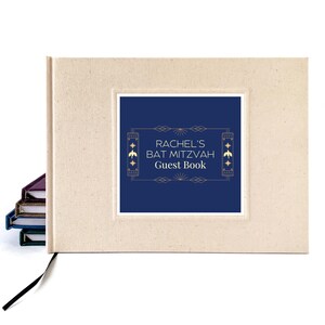 May include: A beige guest book with a navy blue inset featuring the text "RACHEL'S BAT MITZVAH Guest Book" in gold lettering. The book has a black ribbon bookmark and a stack of books is visible in the background.