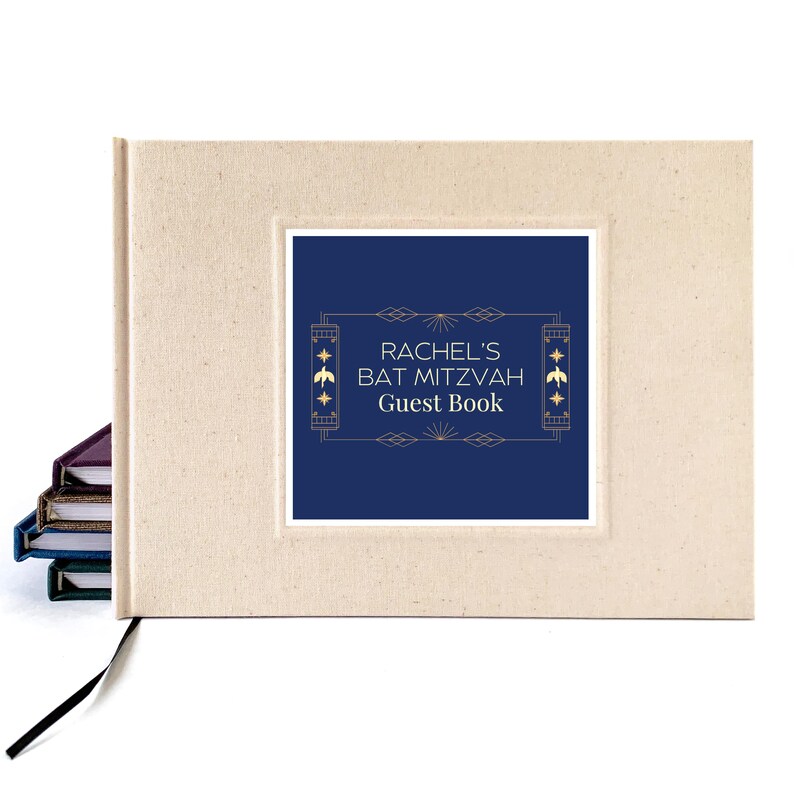 May include: A beige guest book with a navy blue inset featuring the text "RACHEL'S BAT MITZVAH Guest Book" in gold lettering. The book has a black ribbon bookmark and a stack of books is visible in the background.