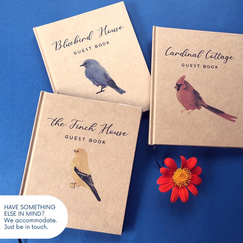 May include: Three tan guest books with bird illustrations and text. One reads Bluebird House, one Cardinal Cottage, and one the Finch House. Each features a different bird: a bluebird, a cardinal, and a finch. A red flower sits near the books.