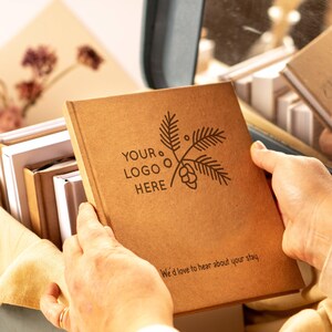 May include: A square brown notebook with a dark brown printed design featuring a pine branch, a pinecone, and the text "YOUR LOGO HERE". The bottom of the cover includes the printed text "We'd love to hear about your stay." The notebook is held open.