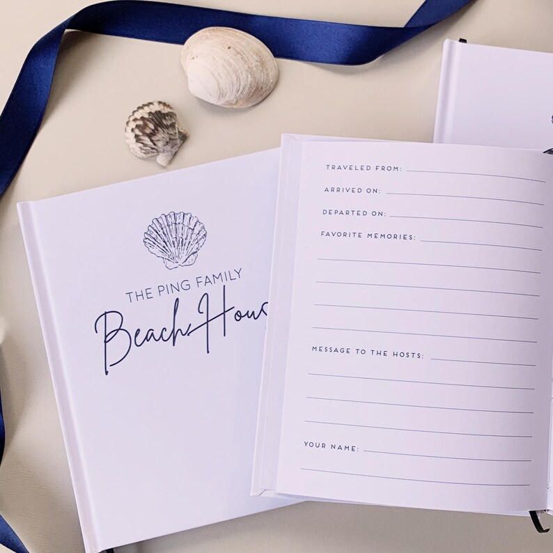 May include: A white guest book with a dark blue shell illustration and the text THE PING FAMILY Beach House on the cover. The open page features printed lines for Traveled From, Arrived On, Departed On, Favourite Memories, Message to the Hosts, and Your Name.