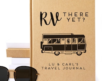 May include: A tan travel journal with the text "RV THERE YET?" and a black illustration of a camper van. The journal also includes the text "LU & CARL'S TRAVEL JOURNAL". Black sunglasses with gold frames are in the foreground.
