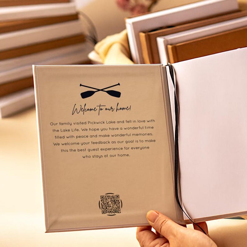 May include: A guest book with a beige cover featuring a black graphic of two crossed oars above the text Welcome to our home! followed by a printed message about Pickwick Lake and a QR code at the bottom. A thin black ribbon bookmark is attached.