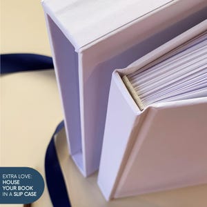 May include: A white book with a matching white slipcase, featuring a visible beige fabric spine binding and white pages. A dark blue ribbon is positioned nearby. The text on the image reads: EXTRA LOVE: HOUSE YOUR BOOK IN A SLIP CASE.