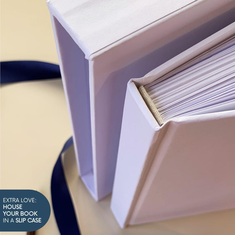May include: A white book with a matching white slipcase, featuring a visible beige fabric spine binding and white pages. A dark blue ribbon is positioned nearby. The text on the image reads: EXTRA LOVE: HOUSE YOUR BOOK IN A SLIP CASE.