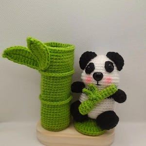 Op de afbeelding: Een gehaakte pandabeer die een groene bamboestok vasthoudt, zittend naast een groene gehaakte bamboe plantenbak op een houten basis.