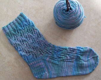 May include: A hand-knitted sock in shades of blue, teal, and lavender, with a decorative openwork pattern. A ball of matching yarn with knitting needles is next to the sock. The sock is a cozy, handmade item.