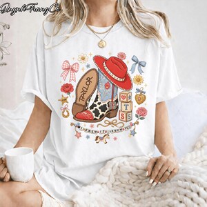Puede incluir: Una camiseta blanca con un estampado gr&aacute;fico de una bota de vaquero con estampado de vaca, un sombrero rojo, el nombre Taylor, el n&uacute;mero 13, bloques de letras, lazos, flores, una herradura, un coraz&oacute;n y una pancarta que dice I KNEW IT I KNEW YOU.