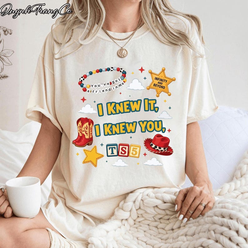 May include: A cream-colored t-shirt featuring a graphic print with the text "I KNEW IT, I KNEW YOU," "TS5," "INFINITY AND BEYOND," and "You've got a friend in me." The design includes a beaded bracelet, a gold star, red cowboy boots, and a red hat.