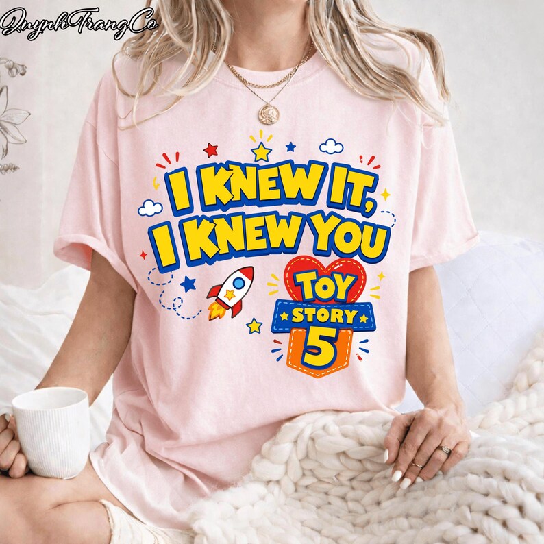 Pu&ograve; includere: Una t-shirt rosa chiaro con il testo I KNEW IT, I KNEW YOU in grassetto giallo con contorni blu. Sotto il testo c'&egrave; la grafica di un razzo e un cuore rosso contenente le parole TOY STORY 5 in giallo e blu.