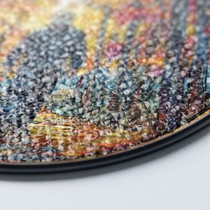 May include: A close-up of a round, colorful, abstract art piece with a black border. The artwork features a mosaic of colors including blue, yellow, orange, and purple. The texture appears to be woven or textured.