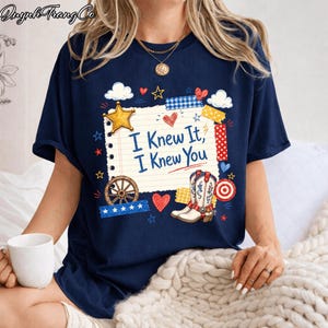 May include: A navy blue t-shirt featuring a graphic of a lined notepad page with the text "I Knew It, I Knew You" in blue. The design includes a yellow star, a pair of western boots, a wagon wheel, red hearts, and various patterned tape strips.