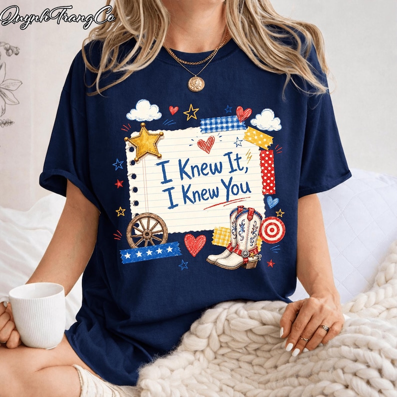 May include: A navy blue t-shirt featuring a graphic of a lined notepad page with the text "I Knew It, I Knew You" in blue. The design includes a yellow star, a pair of western boots, a wagon wheel, red hearts, and various patterned tape strips.