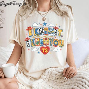 Puede incluir: Una camiseta de manga corta color crema con un estampado gr&aacute;fico colorido que incluye el texto "I KNEW IT, I KNEW YOU" y "TS" dentro de un coraz&oacute;n rojo. El dise&ntilde;o presenta sombreros de vaquero, botas, cactus, estrellas y nubes.