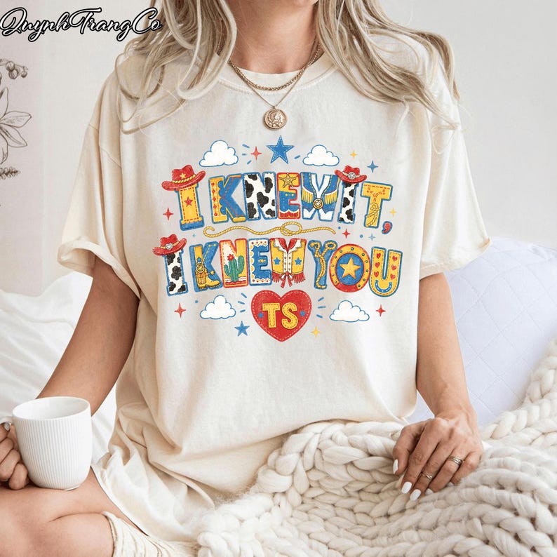 Puede incluir: Una camiseta de manga corta color crema con un estampado gr&aacute;fico colorido que incluye el texto "I KNEW IT, I KNEW YOU" y "TS" dentro de un coraz&oacute;n rojo. El dise&ntilde;o presenta sombreros de vaquero, botas, cactus, estrellas y nubes.