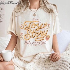 Puede incluir: Una camiseta de manga corta de color crema con un dise&ntilde;o gr&aacute;fico que incluye el texto "it's a Toy Story" escrito en una fuente estilo cuerda, acompa&ntilde;ado de la frase "Baby, just say 'yes'" e ilustraciones de flores, una herradura y un coraz&oacute;n.
