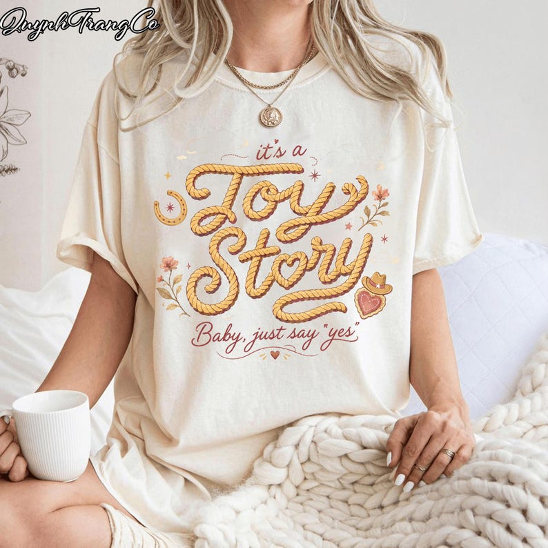 Puede incluir: Una camiseta de manga corta de color crema con un dise&ntilde;o gr&aacute;fico que incluye el texto "it's a Toy Story" escrito en una fuente estilo cuerda, acompa&ntilde;ado de la frase "Baby, just say 'yes'" e ilustraciones de flores, una herradura y un coraz&oacute;n.