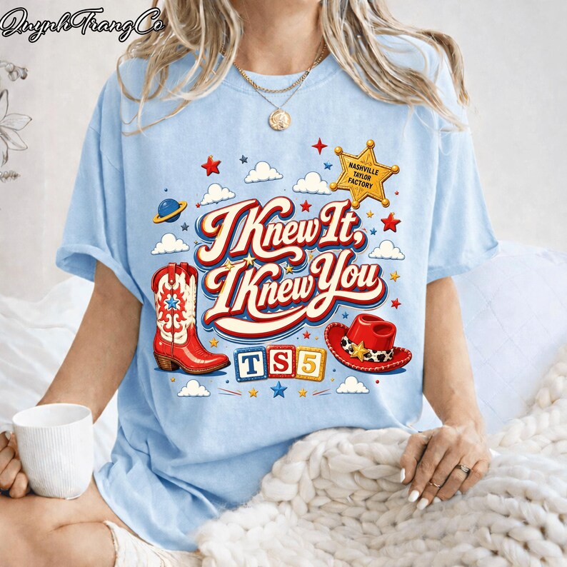 May include: A light blue t-shirt featuring a graphic print with the text I Knew It, I Knew You. The design includes a red cowboy boot, a red cowboy hat, a yellow sheriff star labeled Nashville Taylor Factory, and blocks spelling TS5.